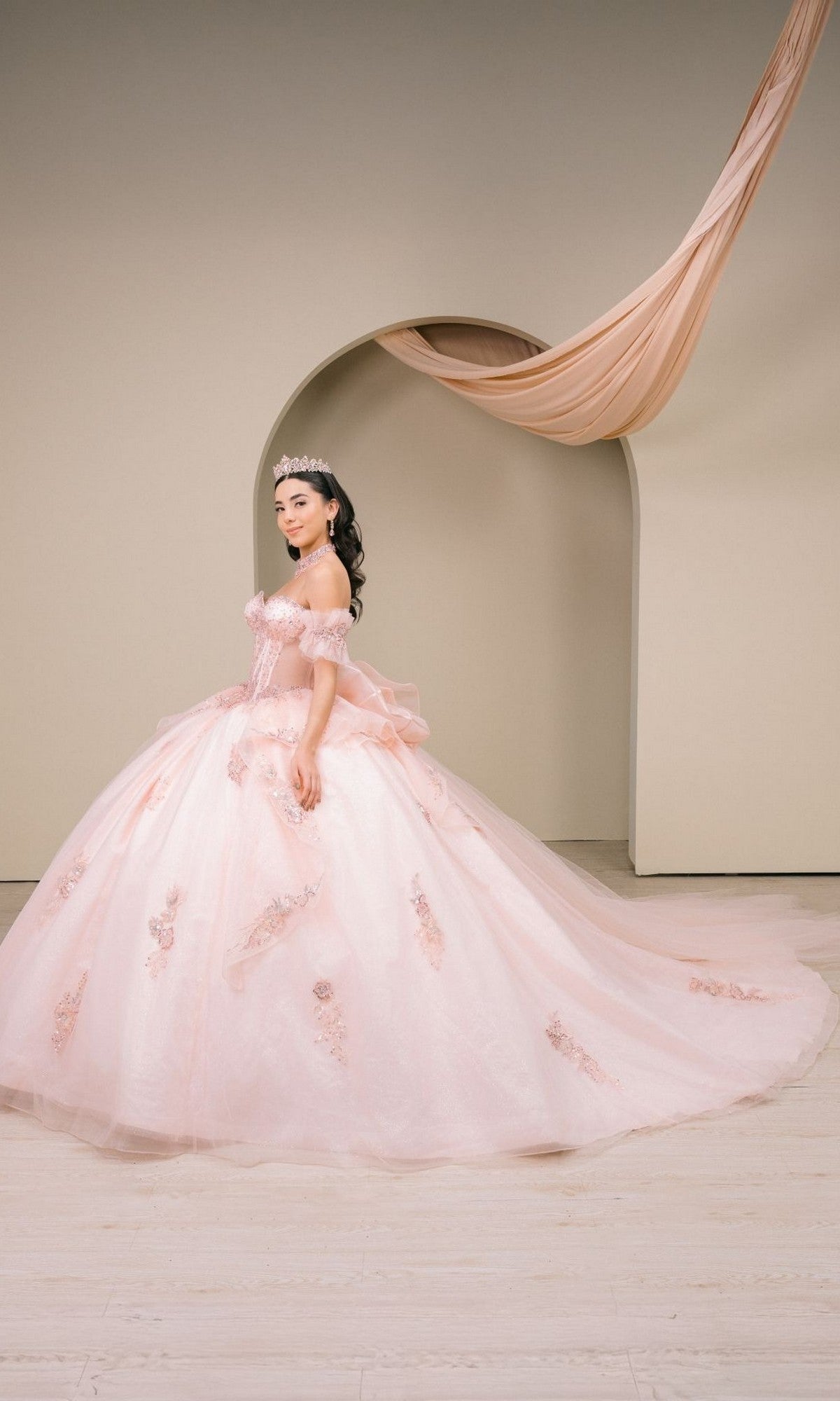 A woman in the Quinceanera Dress 8540JQ by Cinderella Coutureโa blush pink ball gownโpaired with a tiara, stands before a cream-colored arched backdrop adorned with draped fabric.